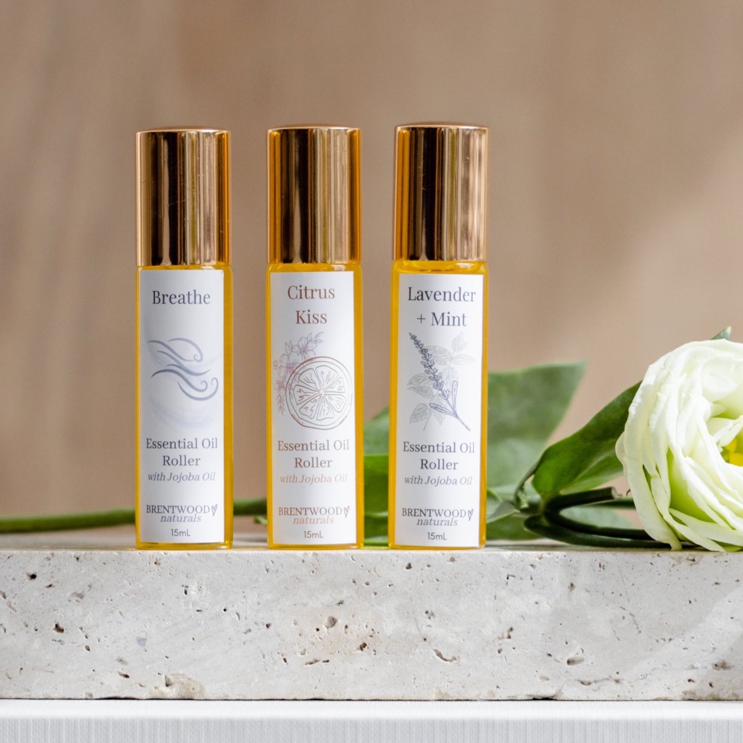 Essential oil rollers in clear glass bottle with shiny gold lid, sitting on a slab on stone with a white rose in the background