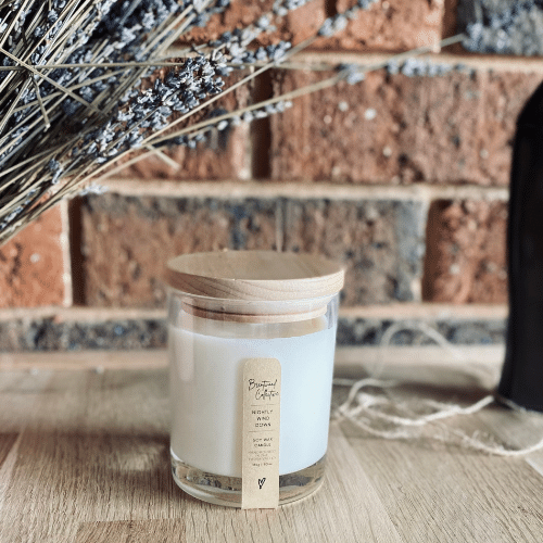 Nightly Wind Down Scented Candle - clear glass jar with natural timber lid & slim/minimal Kraft brown label and black text. Set among dried lavender and curvy female brass statue on natural timber bench top with brick wall in background. 