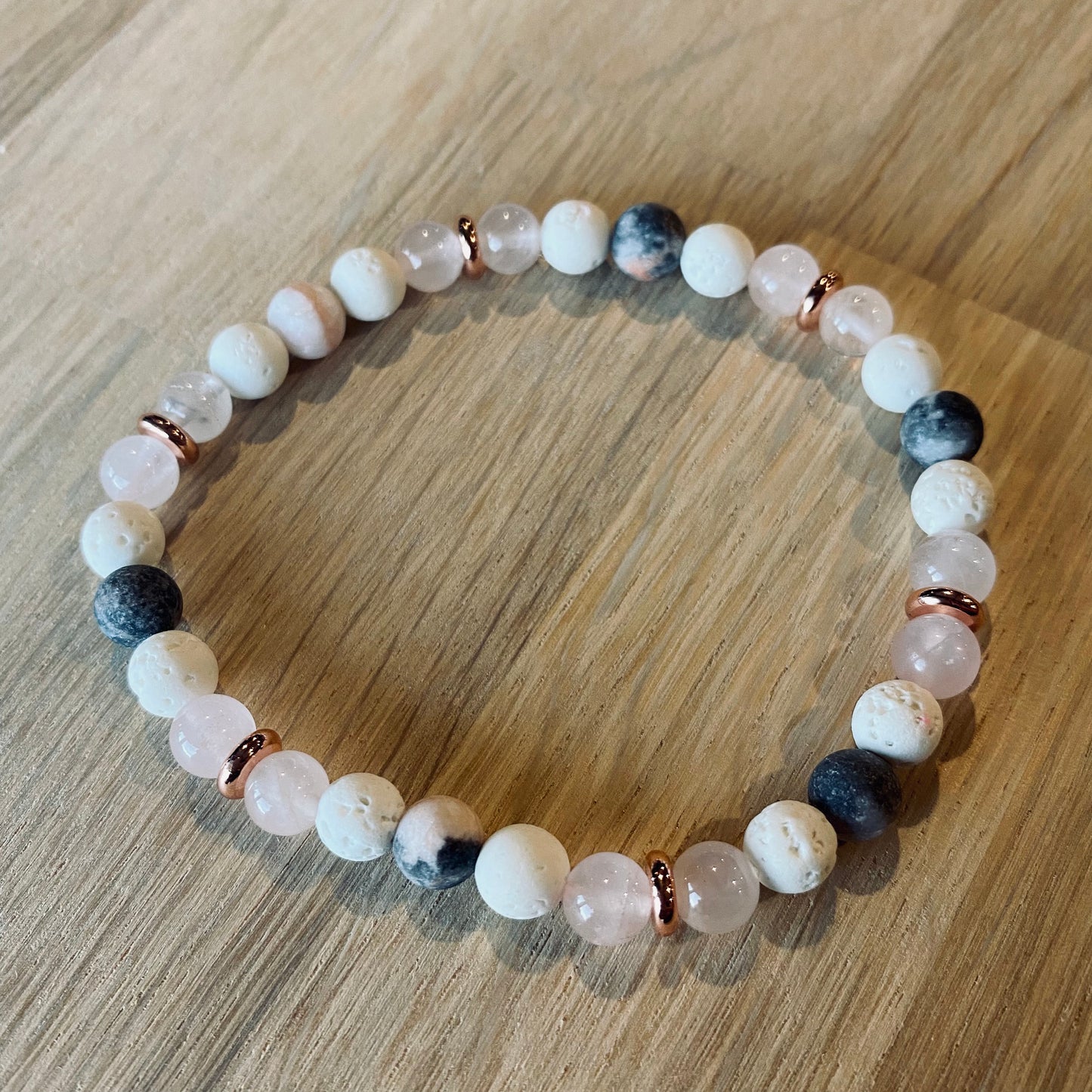 Pink Zebra Jasper Aroma Bracelet - 6mm beaded diffuser bracelet made from a pattern of pink zebra jasper, rose quartz, white lava stone & rose gold disc embellishments. Laid flat on wood grain surface. 