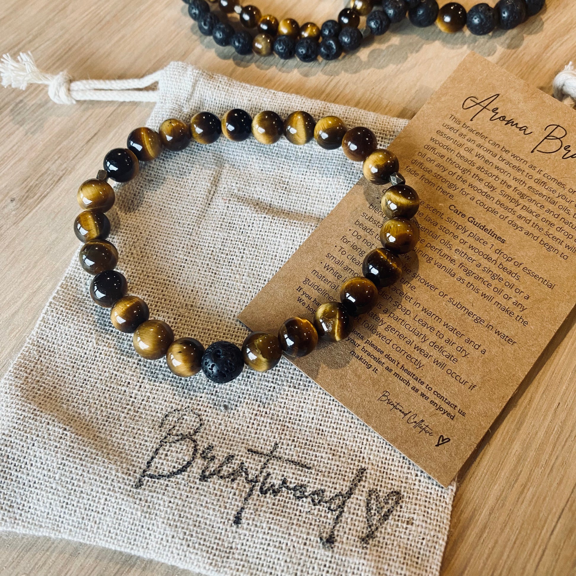 Tiger Eye 6 Aroma Bracelet - 6mm beaded diffuser bracelet made from natural tiger eye gemstone & a single black lava stone bead with antiqued gold faceted embellishments.  Laid on cloth pouch/drawstring bag stamped with “Brentwood” and an aroma bracelet information and care card on kraft brown card.